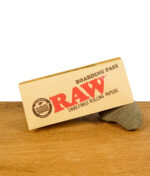 Der geschlossene RAW Boarding Pass auf einem Stein liegend, zeigt das RAW-Logo und den Text 'Unrefined Rolling Papers'.