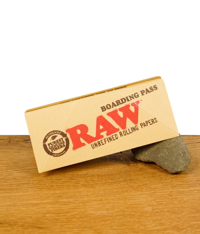 Der geschlossene RAW Boarding Pass auf einem Stein liegend, zeigt das RAW-Logo und den Text 'Unrefined Rolling Papers'.