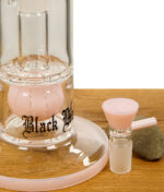 Nahaufnahme der Black Leaf Glasbong mit rosa Kugelperkolator und passendem rosa Glaskopf, dekorativ auf einem Holztisch arrangiert.