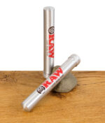 Zwei RAW Aluminium Tubes, eine stehend und eine liegend mit Schraubverschluss, auf Holztisch mit Stein.
