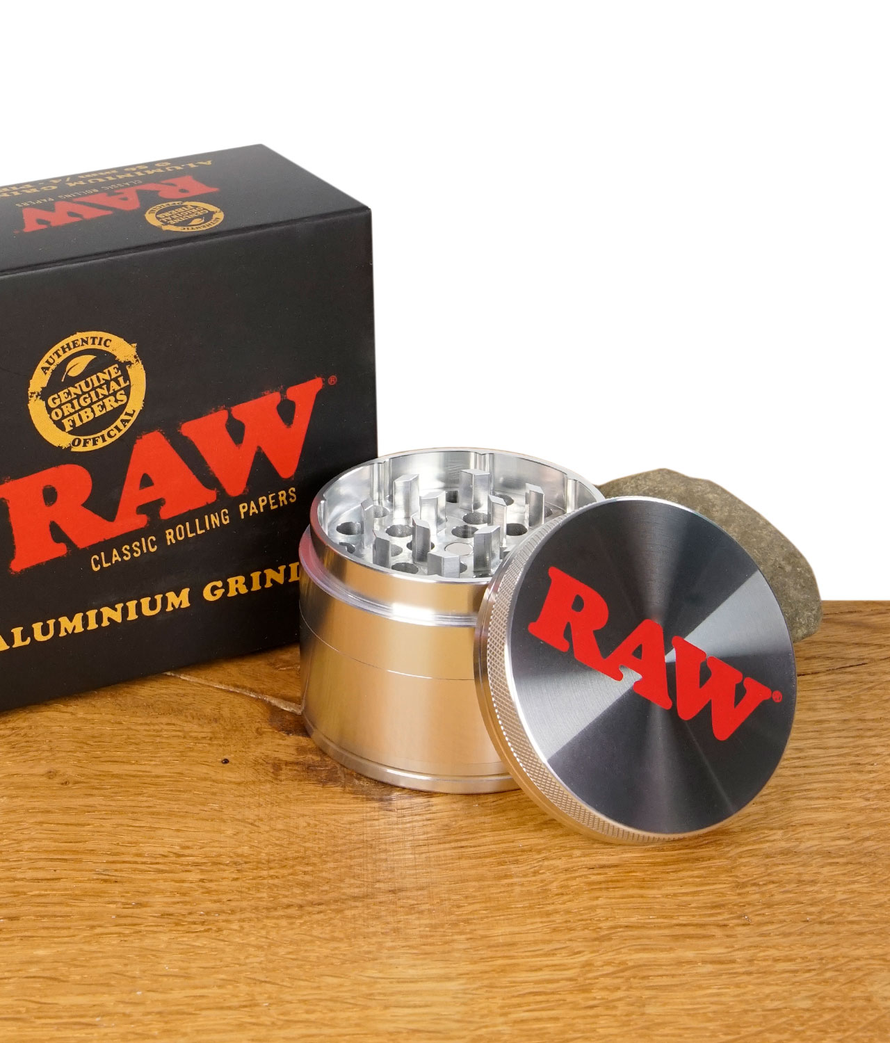 raw-grinder-pollinator-1-jpg Geöffneter RAW Aluminium Grinder mit Verpackung im Hintergrund, Deckel mit rotem RAW-Logo, auf Holztisch mit Stein.