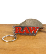 RAW Schlüsselanhänger aus Metall mit rotem Logo und „Classic Rolling Papers“-Schriftzug, liegend auf Holz mit Stein im Hintergrund.