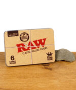 RAW Classic Cone Holder – Metalldose für King Size Cones mit klassischem RAW-Design, platziert auf einem Holztisch mit Stein.