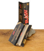 RAW Black Classic Rolling Papers – offene Verpackung mit herausragendem Blättchen, präsentiert auf Holztisch mit Stein.
