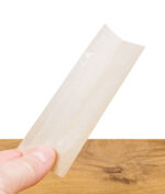 RAW Black Rolling Paper – ultradünnes, ungebleichtes Zigarettenpapier in Nahaufnahme, gehalten zwischen Daumen und Finger vor Holztisch-Hintergrund.