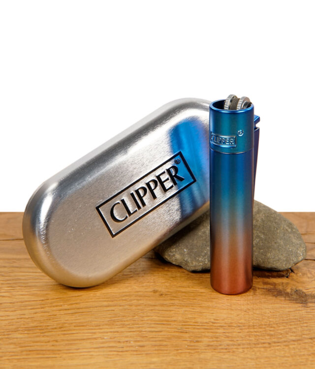 Clipper Metallfeuerzeug im Dawn Gradient Look in Blau-Rot-Farbverlauf vor silberner Clipper-Metallbox auf Holzuntergrund