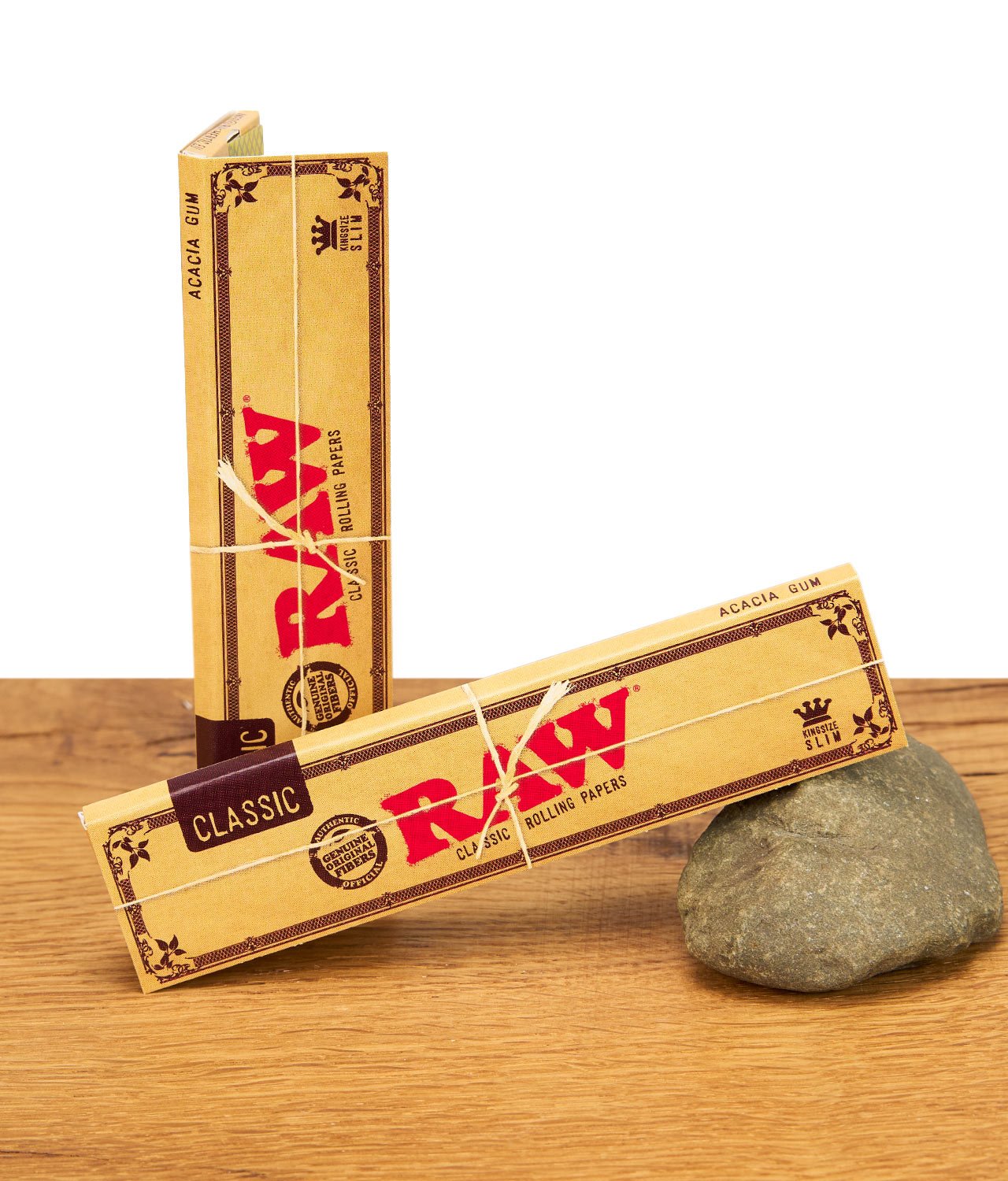 raw-classic-king-size-slim-papers-ungebleicht-jpg Zwei Packungen RAW Classic Kingsize Slim Rolling Papers, mit Naturfaserband umwickelt, auf einem Stein liegend.