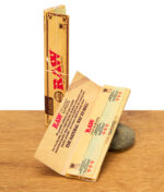 Geöffnete RAW Classic Kingsize Slim Packung mit einem einzelnen Rolling Paper, stehend auf einem Stein.
