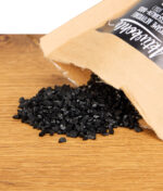 Aufgerissener Beutel mit Purize Aktivkohle; schwarze Aktivkohlekörner aus Kokosnussschale sind auf dem Holztisch verstreut.