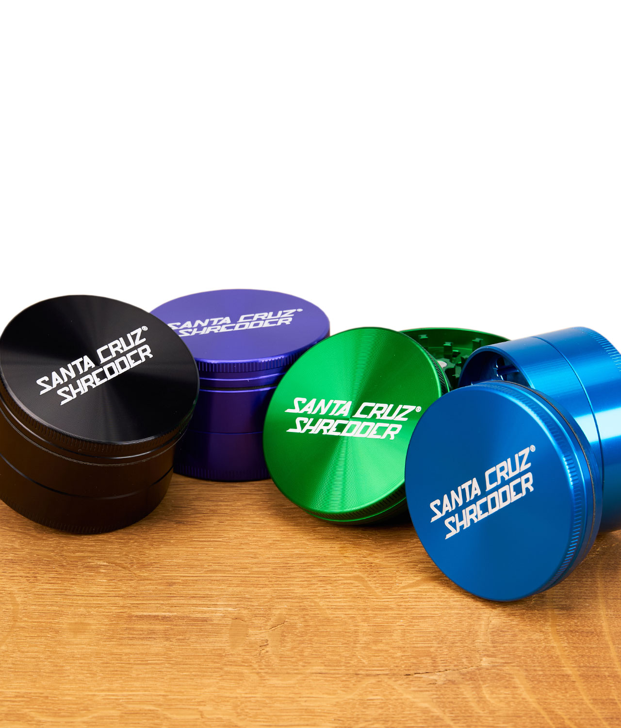 santa-cruz-shredder-3-teilig-gruppenbild-jpg Vier farbige Santa Cruz Shredder Grinder (Schwarz, Lila, Grün, Blau) nebeneinander auf Holzfläche