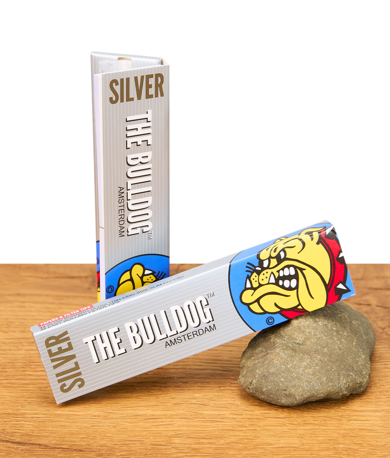 the-bulldog-silber-papers-ks-tips-jpg Zwei Packungen The Bulldog Silver King Size Papers + Tips auf Holztisch, eine stehend und eine auf einem Stein liegend