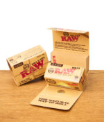 Zwei geöffnete RAW Connoisseur Rolls & Tips Classic Boxen mit sichtbarem Innenleben und Text, daneben eine geschlossene Box, alles auf einem Holztisch.