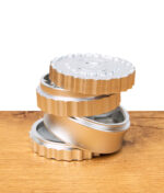 Stapel aus vier silber-goldenen Aluminium-Grinder-Teilen auf Holzuntergrund, Zahnung und Struktur der Pfeffermühle deutlich sichtbar.