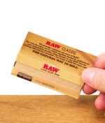 RAW Classic 1 1/4 Size Papers offen auf Holztisch, Person hält Papierpackung in der Hand – natürliche, ungebleichte Blättchen zum Drehen von Zigaretten