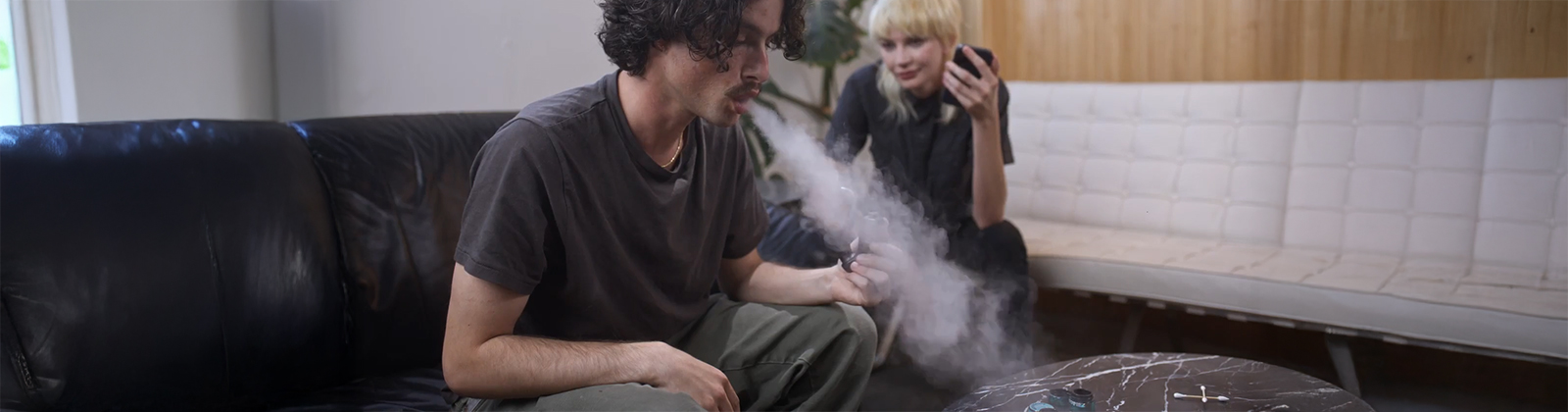Junger Mann atmet Wolke aus Cannabis Vaporizer mit viel Dampf aus. Frau im Hintergrund schaut zu, in gemütlicher Atmosphäre