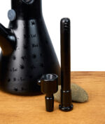 Schwarzes Bong-Zubehörset mit Chillum und Köpfchen neben schwarzer Black Leaf Beakerbong auf Holztisch.