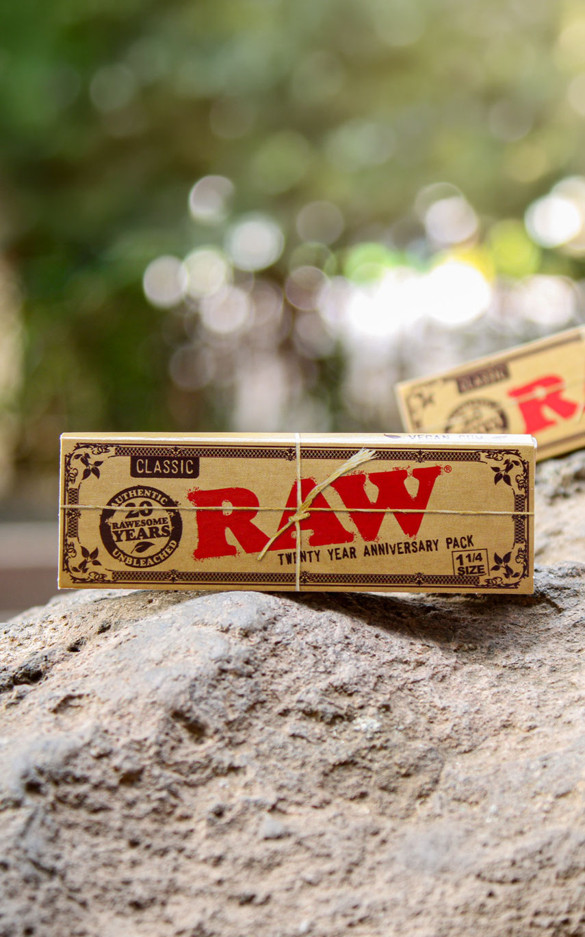 RAW Papers kaufen. RAW Rolling Papers Classic Packung auf einem Stein dem klassischen roten Logo. Weitere Packung im Hintergrund zu sehen vor verschwommenem Hintergrund & Bäumen.