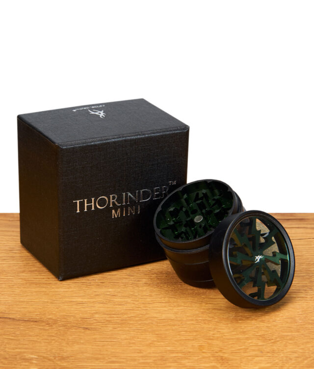 Geöffneter schwarzer Kräutermühle mit grünem Mahlwerk und passender schwarzer Geschenkbox auf Holztisch.