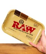 Hand hält ein beige‑farbenes RAW Drehunterlage-Tablett mit rotem RAW Logo und Text „Natural Unrefined Rolling Papers“ über einer Holzoberfläche, Produktfoto schräg von vorn.