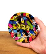 Hand hält einen runden Clipper Aschenbecher mit buntem Feuerzeug-Allover-Design und gelbem Clipper-Logo über einer Holzoberfläche vor weißem Hintergrund.