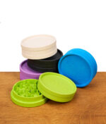 Fünf Santa Cruz Kunststoff-Grinder in verschiedenen Farben stehen auf einer Holzoberfläche vor weißem Hintergrund: Creme, Schwarz, Neongrün, Blau und Violett, teilweise mit geöffnetem Deckel und Zähne innen sichtbar.