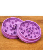 Zwei violette Santa Cruz Grinder mit Kunststoffriffeln stapelweise nebeneinander liegen auf einer Holzoberfläche, beide Hälften zeigen die innere Zahnstruktur zum Zerkleinern.