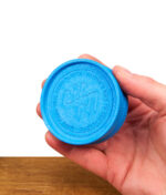 Hand hält einen geöffneten blauen Santa Cruz Grinder mit Drehaufdruck, sichtbar die Zahnstruktur im Inneren der beiden Hälften über einer Holzoberfläche vor weißem Hintergrund.