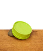 Santa Cruz Grinder in Neongrün mit geschlossenem Deckel, auf dem ein geriffelter Rand sichtbar ist, steht auf einer Holzoberfläche vor weißem Hintergrund, daneben ein grauer Stein.