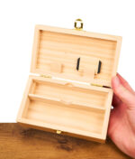 Hand hält eine geöffnete Holzbox mit geteiltem Innenfach und kleinen Haltern im Deckel über einer Holzoberfläche vor weißem Hintergrund.