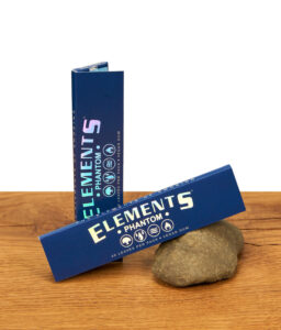 Zwei dunkelblaue Elements Phantom King Size Slim Packungen auf Holzgrund, eine stehend und eine an einen Stein gelehnt.