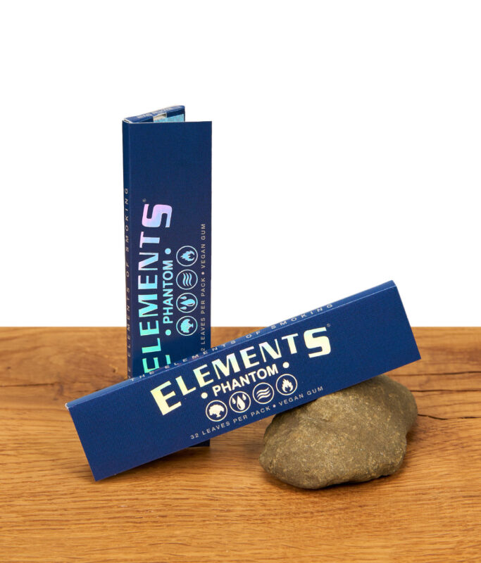 Zwei dunkelblaue Elements Phantom King Size Slim Packungen auf Holzgrund, eine stehend und eine an einen Stein gelehnt.