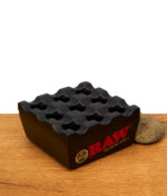 RAW Regal Ashtray Black, windresistenter Aschenbecher aus schwerem Metall in Mattschwarz mit gitterartigem Deckel auf Holztisch.