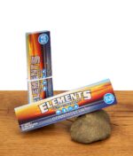 Zwei Packungen Elements Connoisseur King Size Slim Papers mit Tips auf Holzgrund, eine Packung lehnt an einem Naturstein.
