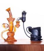 Orangene Premium Bong mit Murmel steht auf Holz mit dem schwarzen Puffco Peak Pro. Sie sind mit dem neuen Kabel des Peak Pro Link verbunden. Weißer Hintergrund.