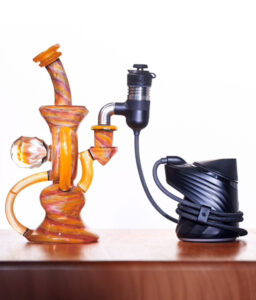 Orangene Premium Bong mit Murmel steht auf Holz mit dem schwarzen Puffco Peak Pro. Sie sind mit dem neuen Kabel des Peak Pro Link verbunden. Weißer Hintergrund.