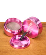 Geöffneter Elements Pink Grinder 4-teilig aus eloxiertem Aluminium. Sichtbares Metallsieb und schwarzer Reinigungspinsel.