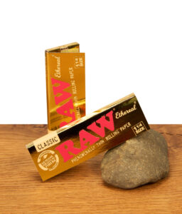 Zwei Packungen RAW Ethereal 1 1/4 Size Papers in glänzender Gold-Optik auf Holzoberfläche mit Naturstein-Akzent.