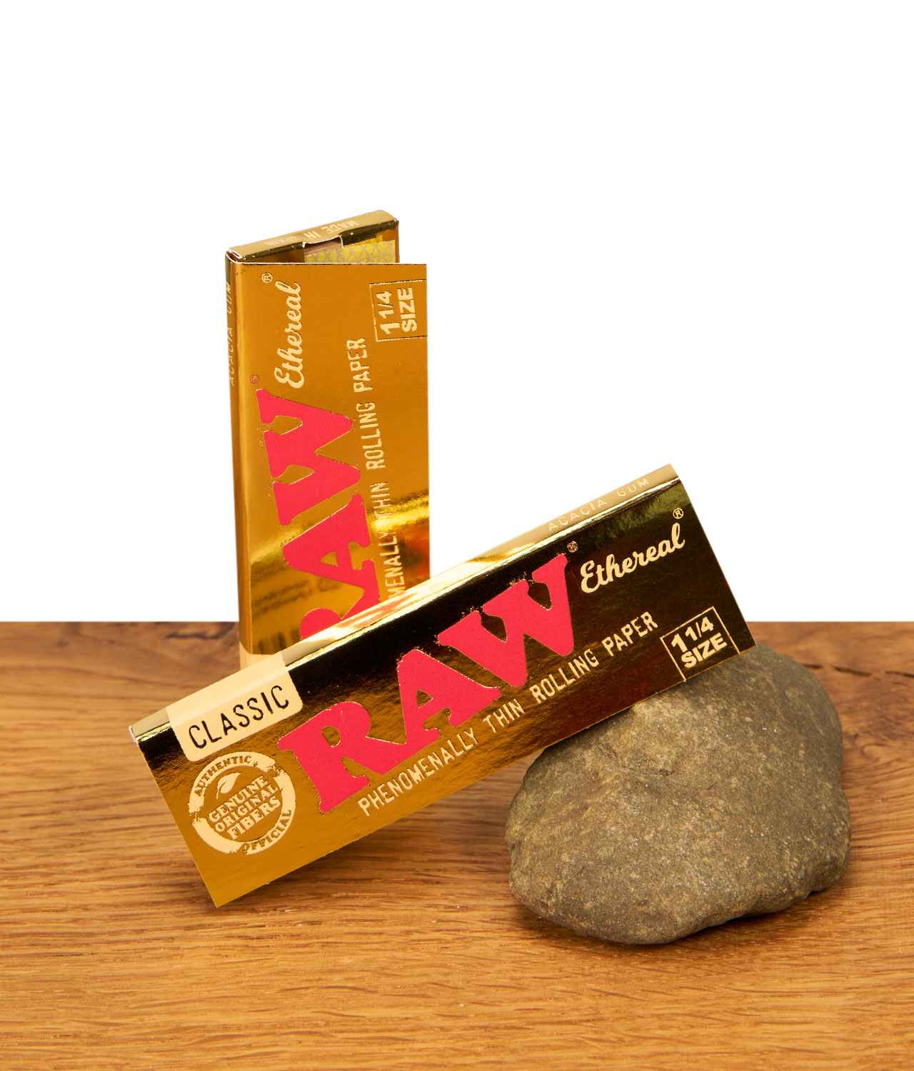 raw-ethereal-ein-ein-viertel-size-paper-jpg Zwei Packungen RAW Ethereal 1 1/4 Size Papers in glänzender Gold-Optik auf Holzoberfläche mit Naturstein-Akzent.