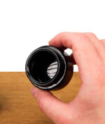 Hand hält eine kleine, geöffnete schwarze Weithalsdose aus Glas über einer Holzoberfläche vor weißem Hintergrund, das Innengewinde ist gut erkennbar.