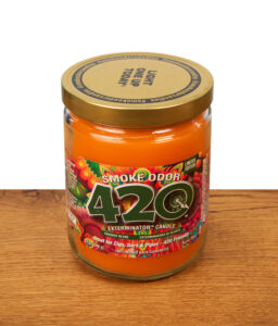 Geschlossene Smoke Odor 420 Exterminator Candle im Glas mit goldenem Deckel auf Holz, orangefarbenes Wachs und farbiges Etikett.