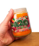 Offene Smoke Odor 420 Exterminator Candle im Glas, in der Hand gehalten, orangefarbenes Wachs und farbiges Etikett über Holz.
