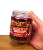 Die 13oz Smoke Odor Cherry Bomb Kerze wird in der Hand gehalten, um die Größe des Glases und das farbenfrohe Design zu verdeutlichen.
