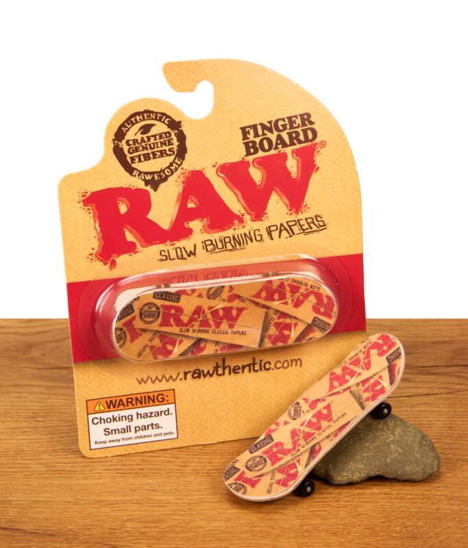 Verkaufsverpackung des RAW Finger Boards mit einem zweiten, ausgepackten Board, das dekorativ an einem Stein auf einer Holzoberfläche lehnt.