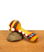 Seitenansicht der bunten B1 Calvo Screen Spoon Pipe mit orange-gelben Streifen und dekorativen weißen Glasnoppen, präsentiert auf einem kleinen Stein.