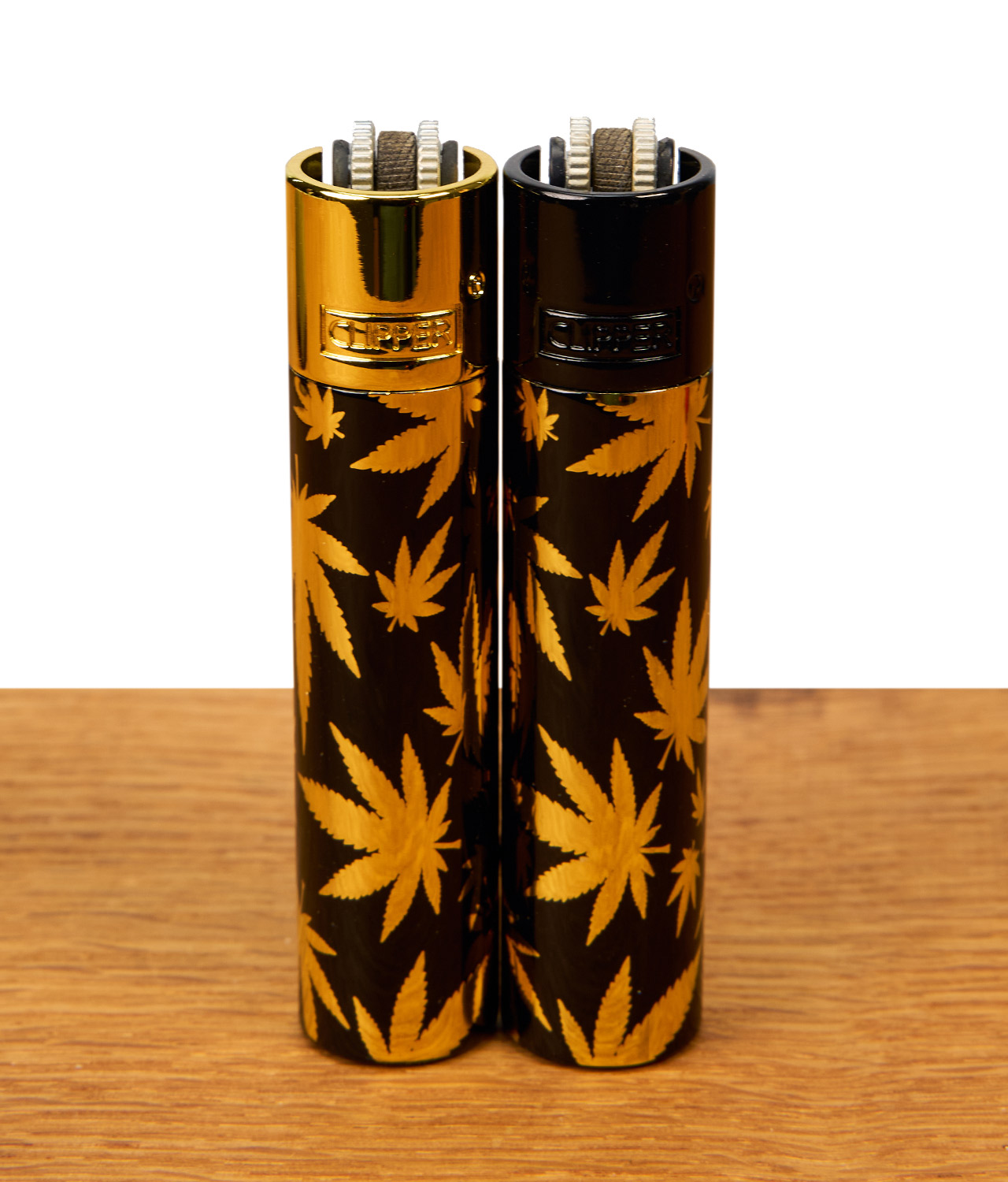 clipper-metall-leaves-gold-jpg Zwei Clipper Metall Leaves Gold Feuerzeuge auf Holz. Schwarzes Metalldesign mit goldenen Hanfblättern, mit schwarzer und goldener Kappe.