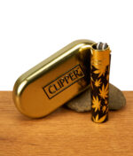 Clipper Metall Leaves Gold Feuerzeug mit goldener Kappe neben goldenem Clipper-Etui auf Holz. Schwarzes Metall mit goldenem Hanfblatt-Motiv.