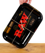 Hand hält das RAW Black Rolling Tray Classic, gebunden mit Naturband, leicht schräg über einem Holztisch.