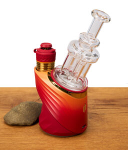 Das Puffco Peak Quadra Top von MJ Arsenal einsatzbereit montiert auf einem rot-orangefarbenen Vaporizer neben einem kleinen Naturstein.