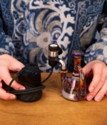 Nutzer mit blau weißem Pullover steht vor einer bunten Glasrig welche mit dem Puffco Peak Pro Link verbunden ist und hält beides in der Hand auf einem Holztisch.