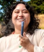 Lifestyle-Aufnahme im Freien, bei der eine Person lächelnd Dampf ausbläst und den saphirblauen Puffco Pivot Sapphire Edition Vaporizer-Pen hält.